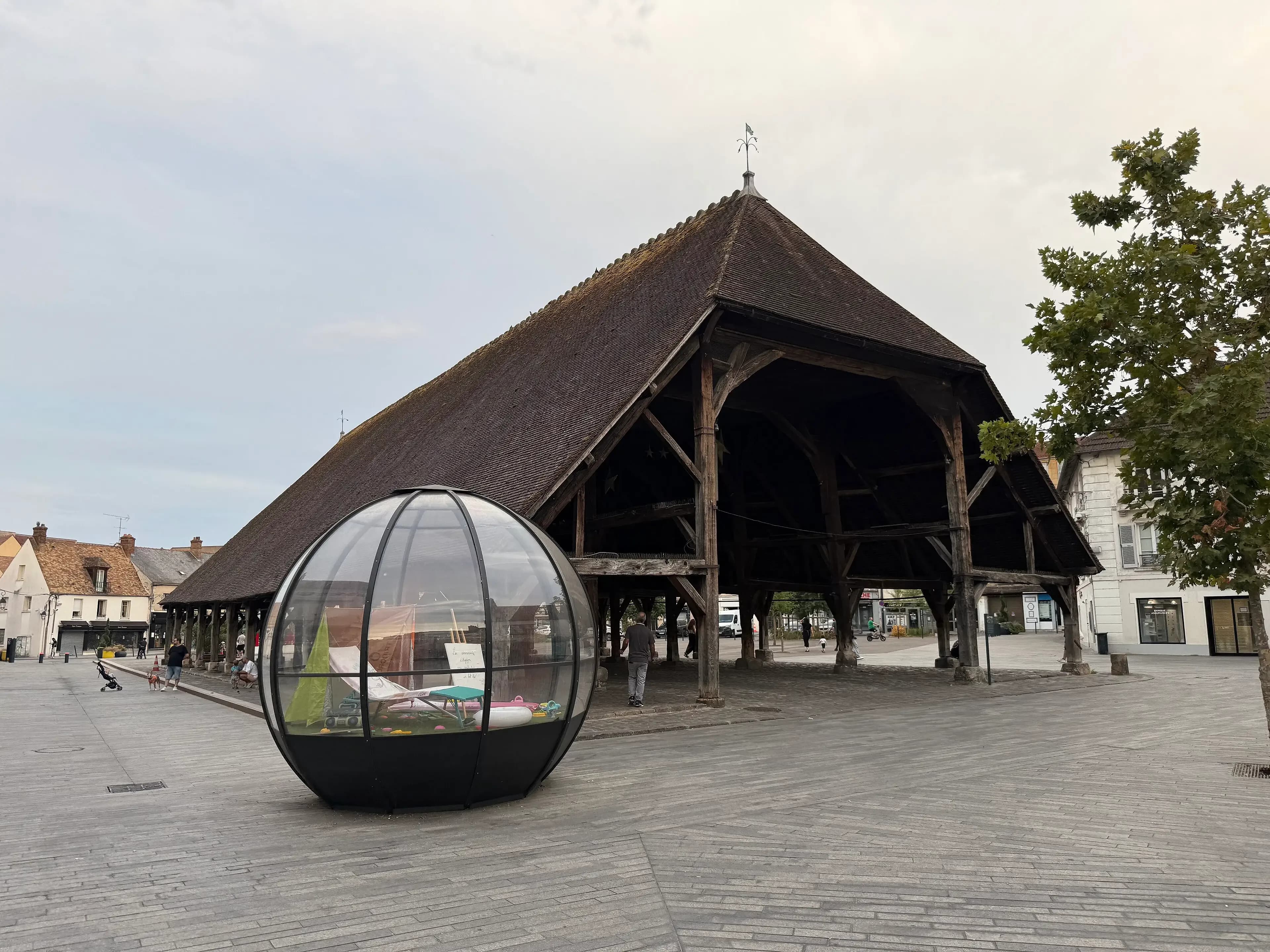 La halle d’Arpajon, vaste toit de tuiles brunes porté par une charpente de bois sur piliers, au bord de la place pavée du centre-ville